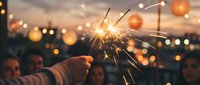 Hand holding a lit sparkler at dusk with blurred city lights and people celebrating in the background, perfect for festive occasions.