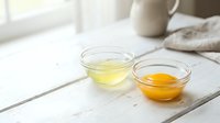 Two glass bowls with separated egg yolk and egg white on rustic white wooden table in bright kitchen setting for cooking and baking preparation.