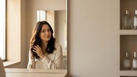 Young Hispanic woman with long dark hair looking at herself in bathroom mirror during morning skincare routine in modern neutral-toned home interior.