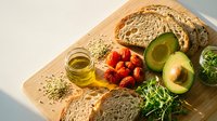Healthy breakfast spread with avocado, whole grain bread, cherry tomatoes, olive oil and fresh greens on wooden cutting board for nutritious meal prep.