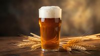 Golden amber beer with creamy foam head in glass mug surrounded by wheat stalks on wooden table with warm bokeh background for brewing and pub concepts.