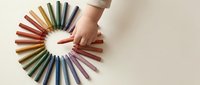 Child hand reaching for colorful wooden crayons arranged in circle on neutral background. Creative art supplies for kids education and drawing activities.
