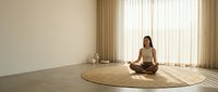 Woman in lotus position meditating on round jute rug in minimalist room with natural light, suitable for wellness, yoga, mindfulness and relaxation content.