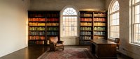 Elegant home library with floor-to-ceiling bookshelves, vintage leather armchair, wooden desk and warm natural lighting from arched windows.