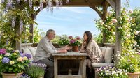 Middle-aged Caucasian couple enjoying romantic moment at wooden garden table under pergola surrounded by blooming flowers and lush greenery outdoors.
