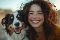 Young Hispanic woman with curly hair and bright smile posing with Australian shepherd dog outdoors during golden hour sunset, expressing joy and companionship.