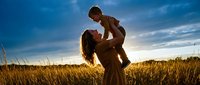 Happy mother lifting toddler son in golden wheat field at sunset creating joyful family bonding moment in rural countryside landscape.