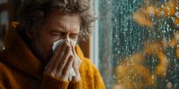 Sick mature man with curly gray hair blowing nose into tissue by rainy window, wearing orange sweater, suffering from cold or flu symptoms during autumn season.