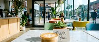 Cozy modern cafe interior with latte art coffee and a thank you note card on marble table, surrounded by green plants and natural light.