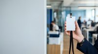 Hand holding blank ID badge with lanyard in modern office environment with blurred colleagues working in background.