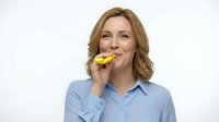 Middle-aged woman with shoulder-length blonde wavy hair and fair complexion wearing light blue button-up shirt, eating yellow apple, smiling at camera against white background.