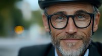 Mature Caucasian man with gray beard wearing glasses and flat cap smiling warmly at camera in outdoor setting with soft bokeh background lighting.