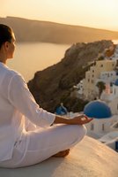 Young woman in white clothing practicing meditation yoga pose overlooking coastal village at golden hour sunset creating peaceful wellness atmosphere.