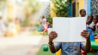 Child holding blank white paper with face covered, wearing patterned blue shirt, outdoors in colorful community setting with blurred figures in background.