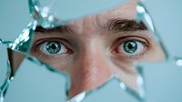Young Caucasian man with striking blue eyes looking through broken glass fragments, conceptual portrait for mental health awareness campaigns.