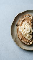 Whole grain toast topped with creamy spread and fresh banana slices arranged on a rustic ceramic plate against a light gray background, perfect for food and nutrition content.