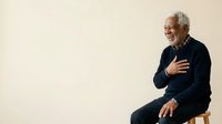 Senior man with gray hair and light skin tone, wearing dark navy sweater, sitting on chair against neutral background, smiling with hand on chest, expressing gratitude or positive emotion for lifestyle and wellness content.