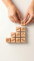 Hands arranging wooden blocks with financial icons including target, growth chart, coins, piggy bank and percentage symbols for business planning.