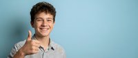 Young Caucasian man with curly brown hair giving thumbs up gesture while smiling confidently against blue background for business and success concepts.