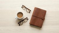 Brown leather journal with coffee cup and reading glasses on wooden desk for business planning and note-taking workspace flatlay composition.