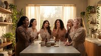 Six diverse women in bathrobes enjoying spa day together in modern wellness center with natural lighting and plants for relaxation retreat.