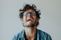 Young Hispanic man with curly hair and glasses wearing denim jacket laughing joyfully with head tilted back against light background, expressing genuine happiness.