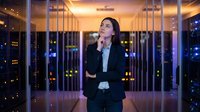 Young woman with long dark hair and East Asian ethnicity, wearing a professional black blazer, standing in a modern server room with illuminated server racks and city lights visible through windows at night, contemplating technology infrastructure.