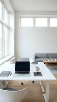 Minimalist modern home office with white desk, open laptop, tablet, coffee mug, and gray sofa in bright natural light.