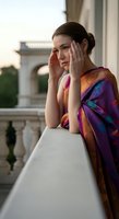 Young Caucasian woman wearing colorful patterned shawl leaning on balcony railing with hands to face, contemplative expression, classical architecture background, suitable for lifestyle and editorial use.