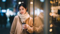 Young Asian woman in beige coat and scarf holding coffee cup on city street at night with bokeh lights background for lifestyle and urban concepts.