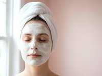 Young woman with white clay facial mask and towel on head relaxing during spa skincare treatment at home with eyes closed for beauty wellness routine.