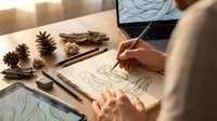 Young woman artist sketching nature drawings at wooden desk with pine cones, colored pencils and laptop in warm sunlit workspace for creative design projects.