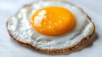 Macro shot of sunny side up fried egg with crispy brown edges and bright orange-yellow yolk on white surface, showing texture and cooking details.