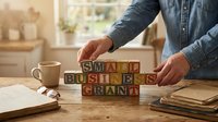 Person arranging wooden letter blocks spelling small business grant on desk with coffee cup and papers in bright office workspace for entrepreneurship.