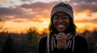 Young African American woman wearing knit hat and scarf smiling warmly at golden hour sunset with dramatic sky and silhouetted trees in background for lifestyle content.