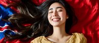 Young Asian woman with flowing dark hair lying on vibrant red and blue fabric, wearing yellow top, expressing joy with genuine smile and closed eyes in relaxed pose.