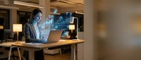 Professional businesswoman analyzing financial data on laptop with holographic charts and graphs in modern office environment during evening work session.