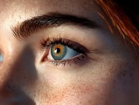 Close-up view of bright blue eye with long eyelashes and natural freckles on fair skin in warm sunlight, showing intricate iris detail and natural beauty.