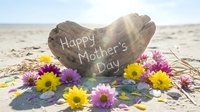 Heart-shaped stone with Happy Mother's Day message surrounded by colorful flowers on sandy beach with sunlight creating warm atmosphere for celebration.