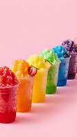 Colorful rainbow shaved ice drinks lined up on pink background, each cup filled with vibrant flavored syrup and crushed ice topped with fresh fruit garnish for summer beverages.