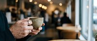 Man holding coffee cup in modern cafe with warm lighting and blurred customers in background creating cozy atmosphere for relaxation and social gathering.