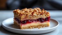 Layered berry crumble bar with cream cheese filling and crunchy streusel topping on white plate, close up view showing texture and layers of sweet dessert.