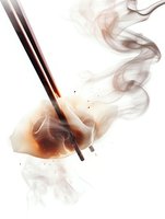 Dramatic close-up of chopsticks dipping into steaming soy sauce with rising smoke swirls and droplets against white background, creating an artistic culinary scene.