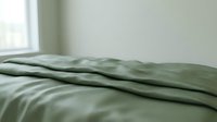 Unmade bed with wrinkled sage green linen sheets and pillowcase in minimalist bedroom interior with natural window light creating peaceful morning atmosphere.