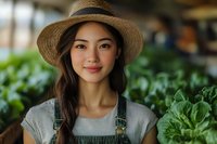 Young Asian female farmer wearing straw hat and denim overalls smiling in vegetable garden greenhouse. Agriculture, farming, gardening, sustainable living.