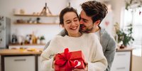 Young couple sharing a joyful moment in cozy kitchen as man surprises woman with red gift box tied with ribbon.