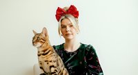 Young woman with blonde hair wearing red bow headpiece and dark patterned sweater holding tabby cat against white background for holiday portraits and festive campaigns.