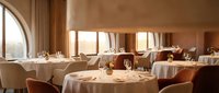 Elegant fine dining restaurant interior with round tables set for service, warm lighting and large windows creating sophisticated upscale atmosphere.