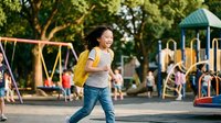Happy Asian elementary school girl with yellow backpack walking at playground during sunny day with colorful equipment and trees in background for education concepts.