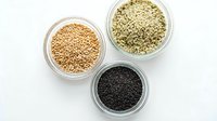 Three glass bowls containing different types of seeds including golden quinoa, hemp seeds and black chia seeds arranged on white background for healthy cooking.